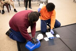 teacher giving CPR instructions to a youth in orange shirt